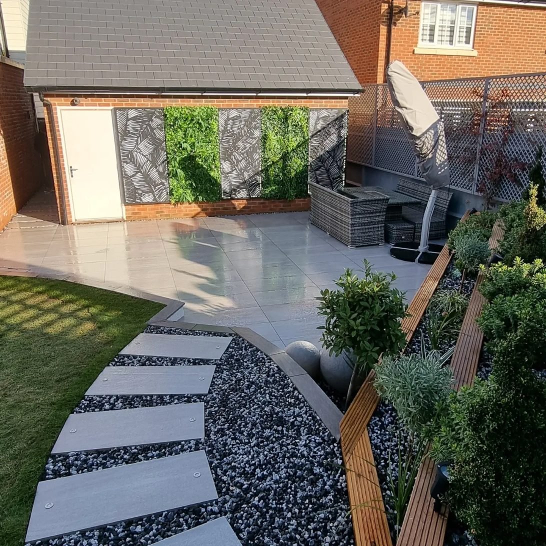 Pale porcelain stepping slabs curving through black stone chippings beside a lawn, with timber raised beds, a porcelain patio and green wall panels between leaf-pattern screens on a garage wall.