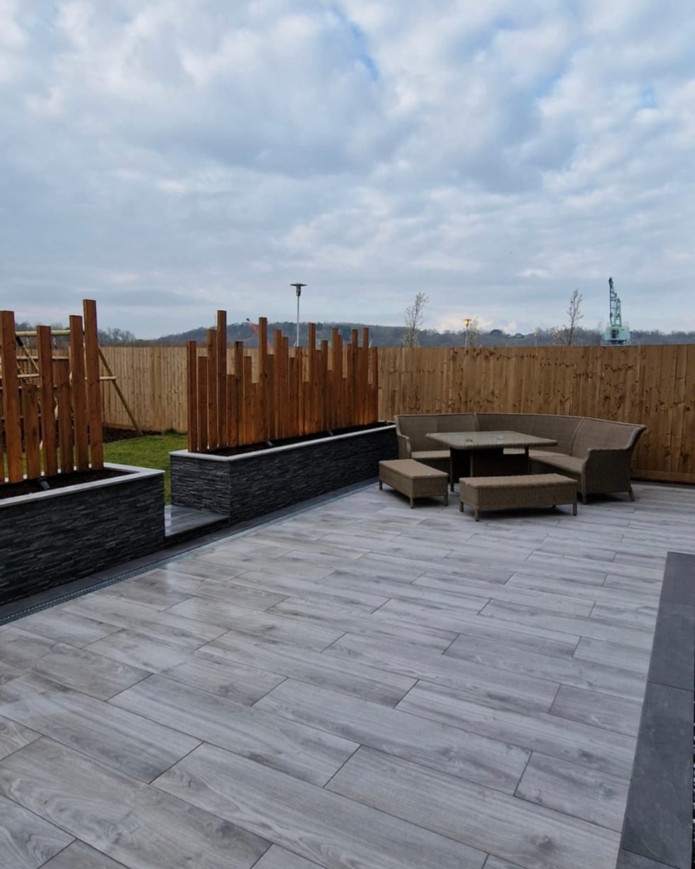 Wood-effect grey porcelain paving with dark stone-clad planters topped by staggered vertical timber posts.