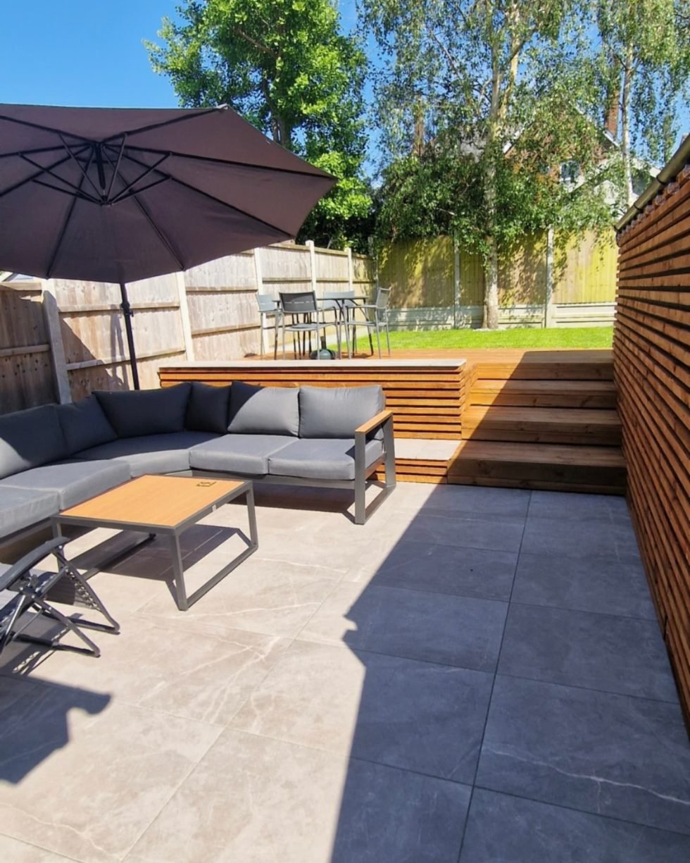 A sunken porcelain terrace with an outdoor sofa, below timber-clad steps and a horizontal slatted timber screen, with a raised dining deck and lawn beyond.