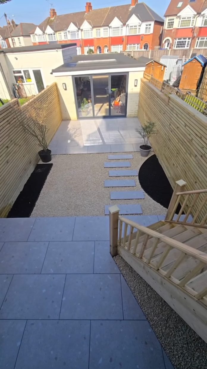 A long terraced garden seen from a raised patio: grey porcelain, timber steps down to a gravel garden with stepping slabs, slatted fences and black-mulched beds.