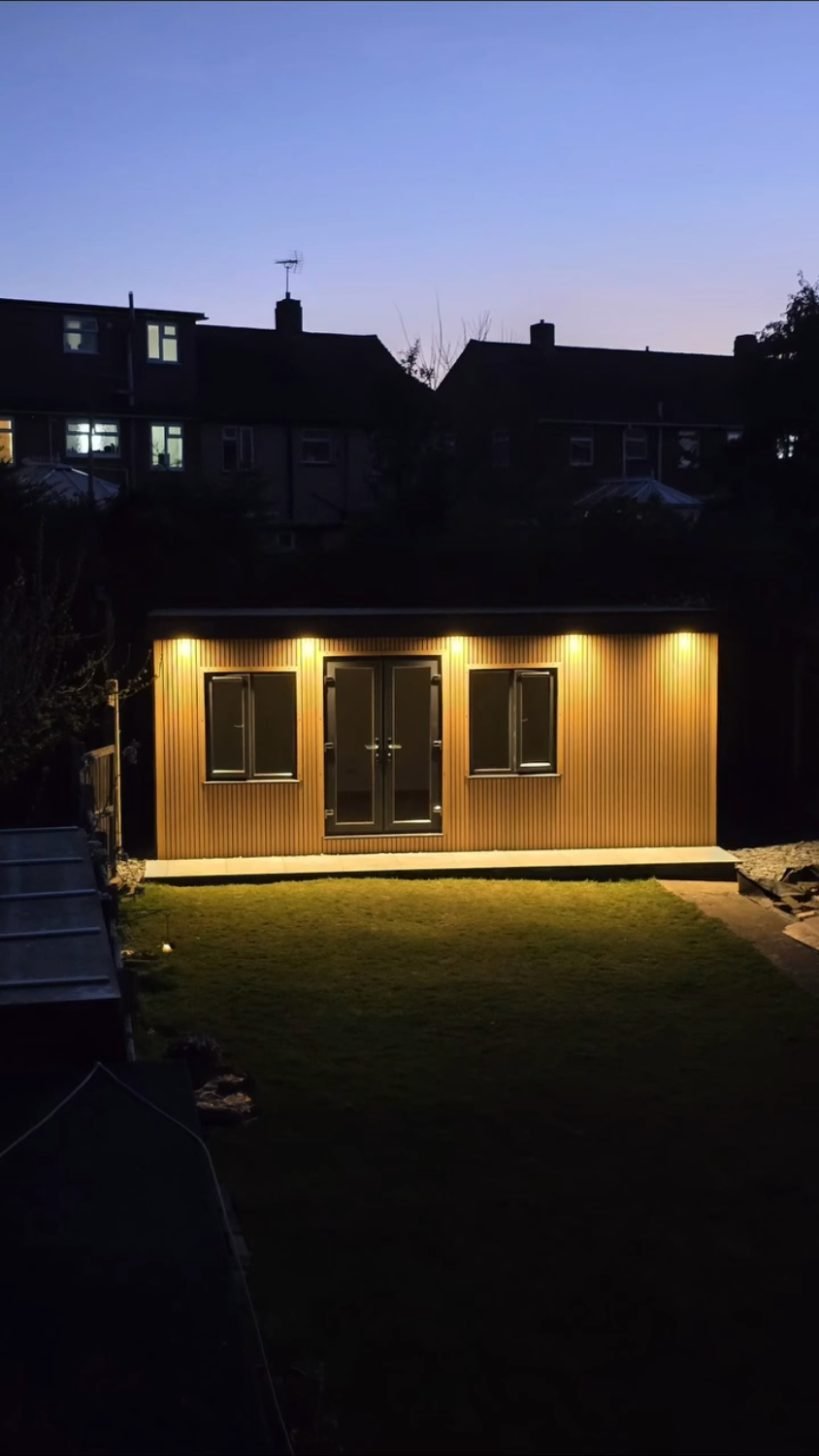 A timber-clad garden room at dusk with soffit lights washing down its slatted front.