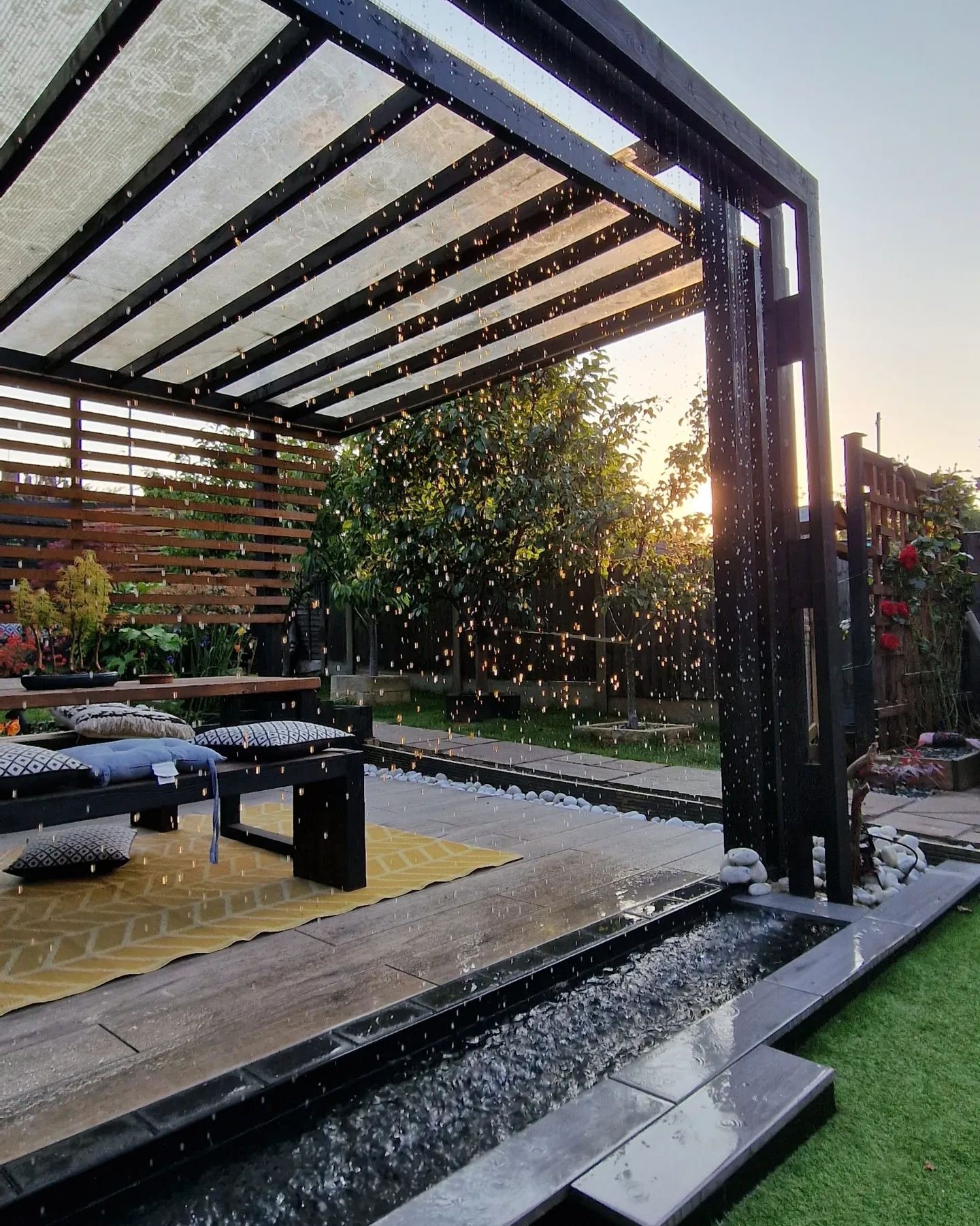 A black timber pergola at sunset. A curtain of water falls from its beam into a pebble-filled rill, catching the low sun, with a bench, cushions and a yellow rug beneath the clear roof.