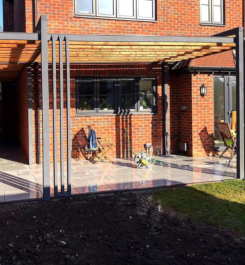 A grey timber pergola across the back of a red-brick house, its posts gathered in bundles of slim uprights, with porcelain paving beneath.