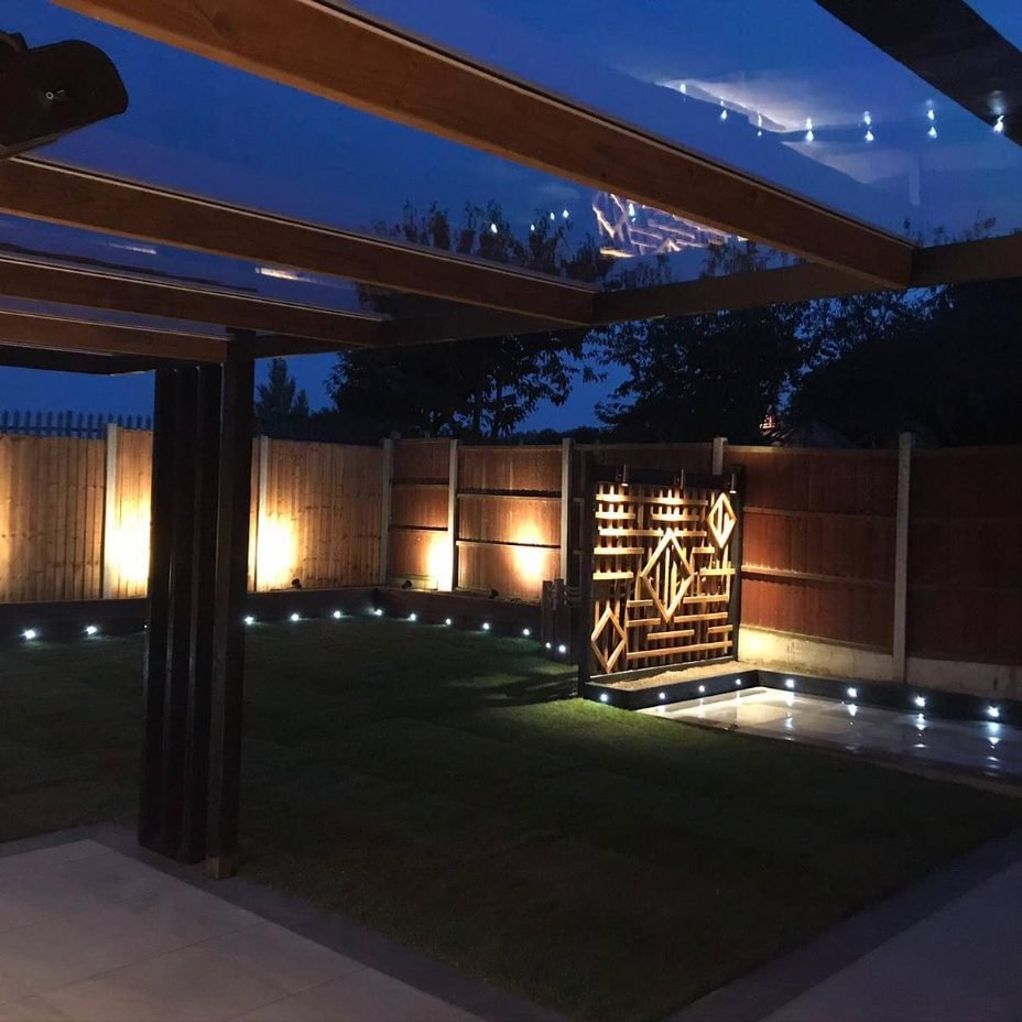 Looking out from under a clear-roofed pergola at night towards a lit timber lattice screen with a diamond pattern, uplit fence panels and lights along the lawn edge.
