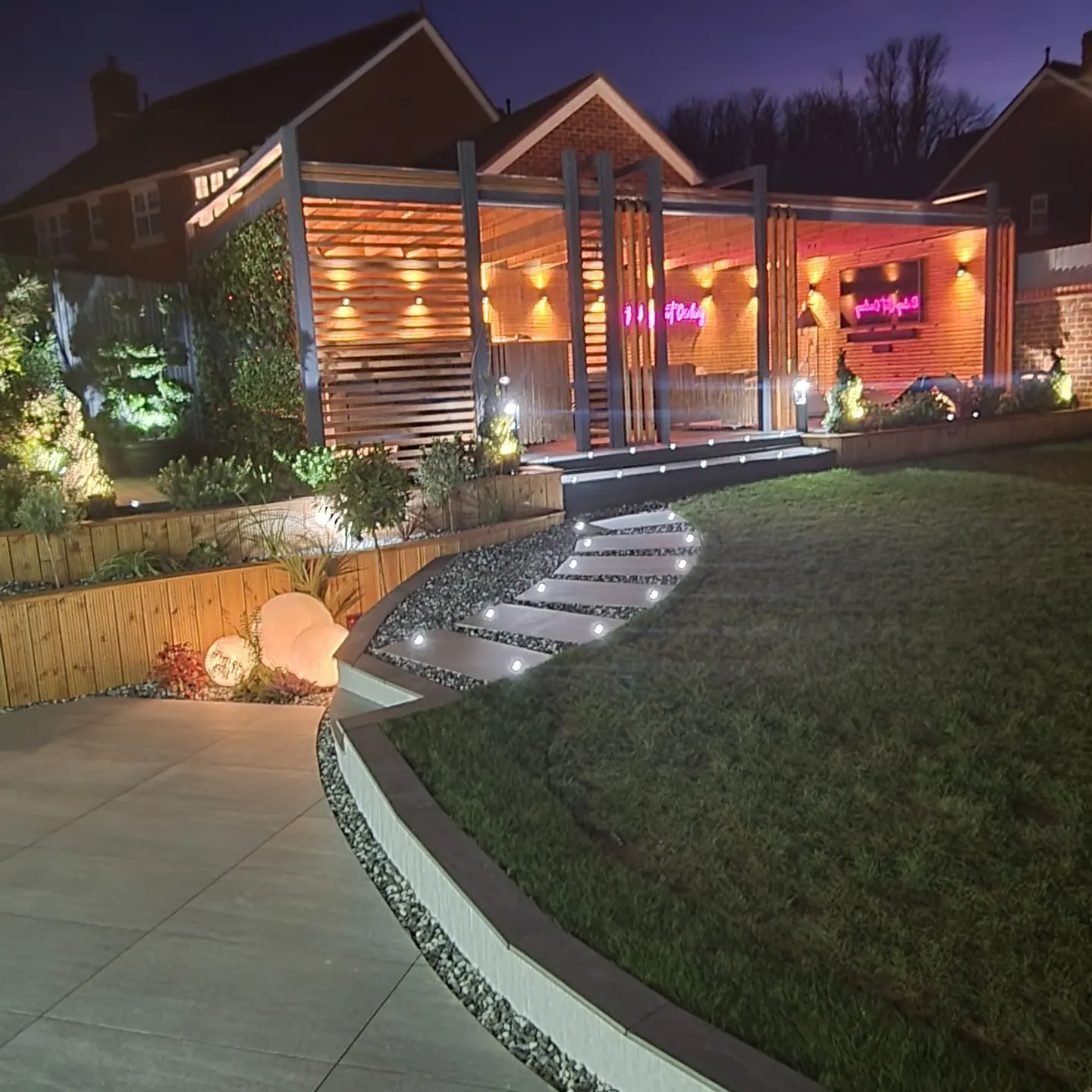 A large pergola at night with slatted timber walls and warm wall lights, reached by lit stepping slabs that curve up beside a lawn and timber raised beds.