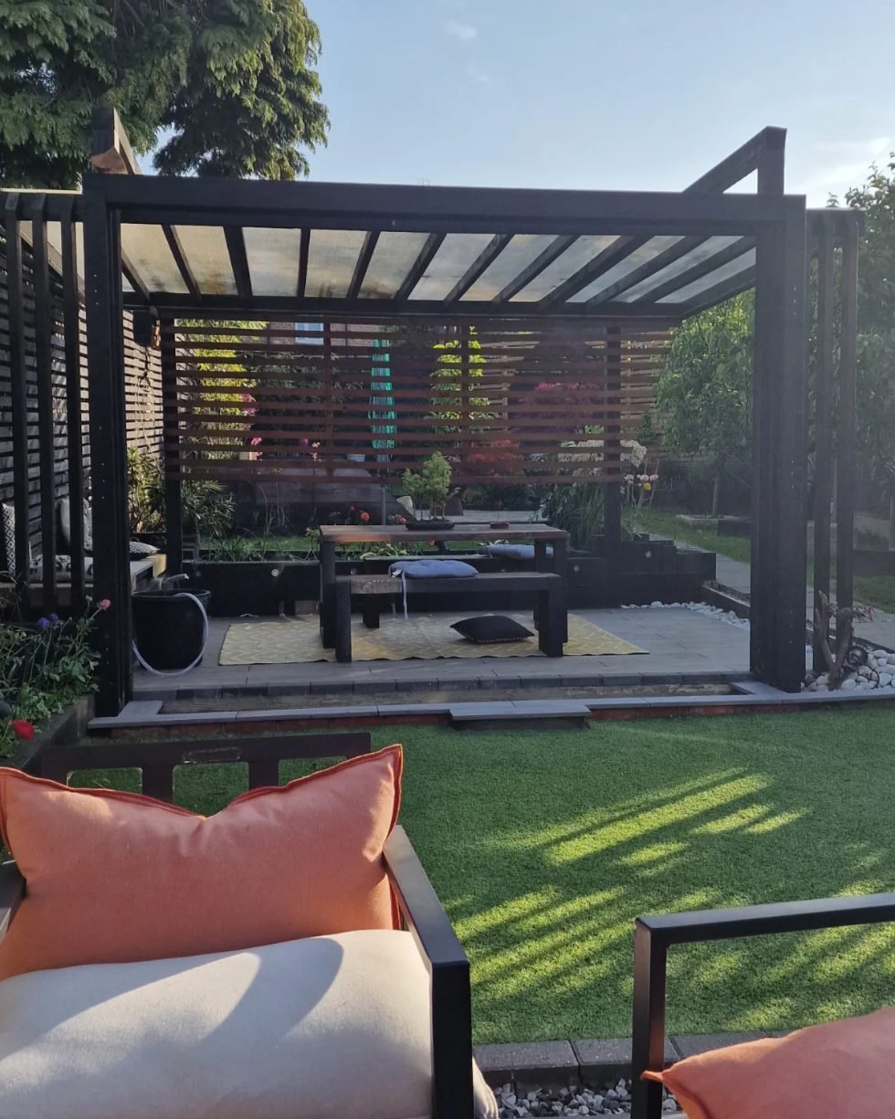 A black timber pergola with a clear roof and a slatted back screen, sheltering a bench and table on a yellow rug, seen across a lawn from a sofa with orange cushions.