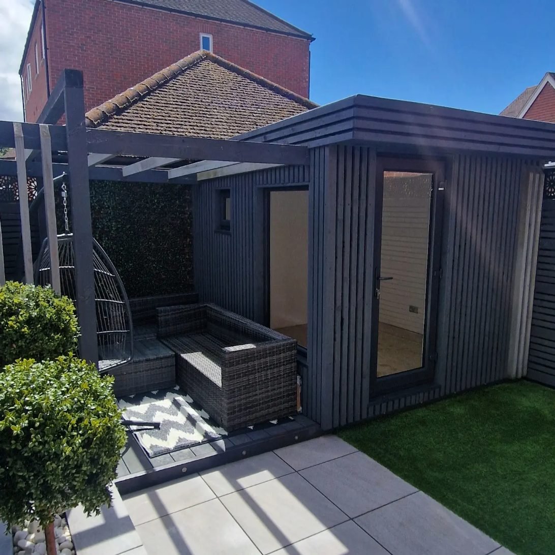 A charcoal timber pergola with a hanging egg chair and rattan seating, joined to a new charcoal-clad garden studio with a glazed door.