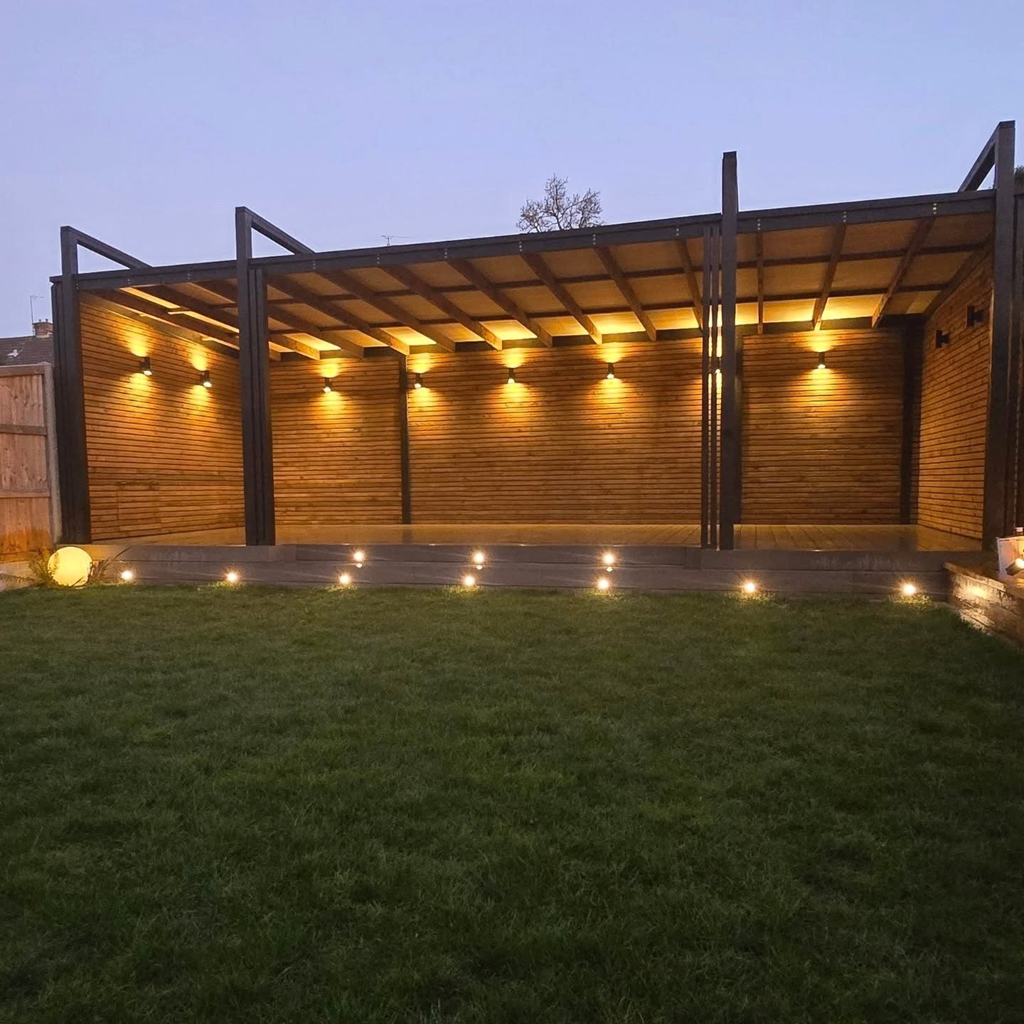 A long timber pergola at dusk, its slatted back wall lit by a row of wall lights, with deck lights along the front edge and lawn in the foreground.