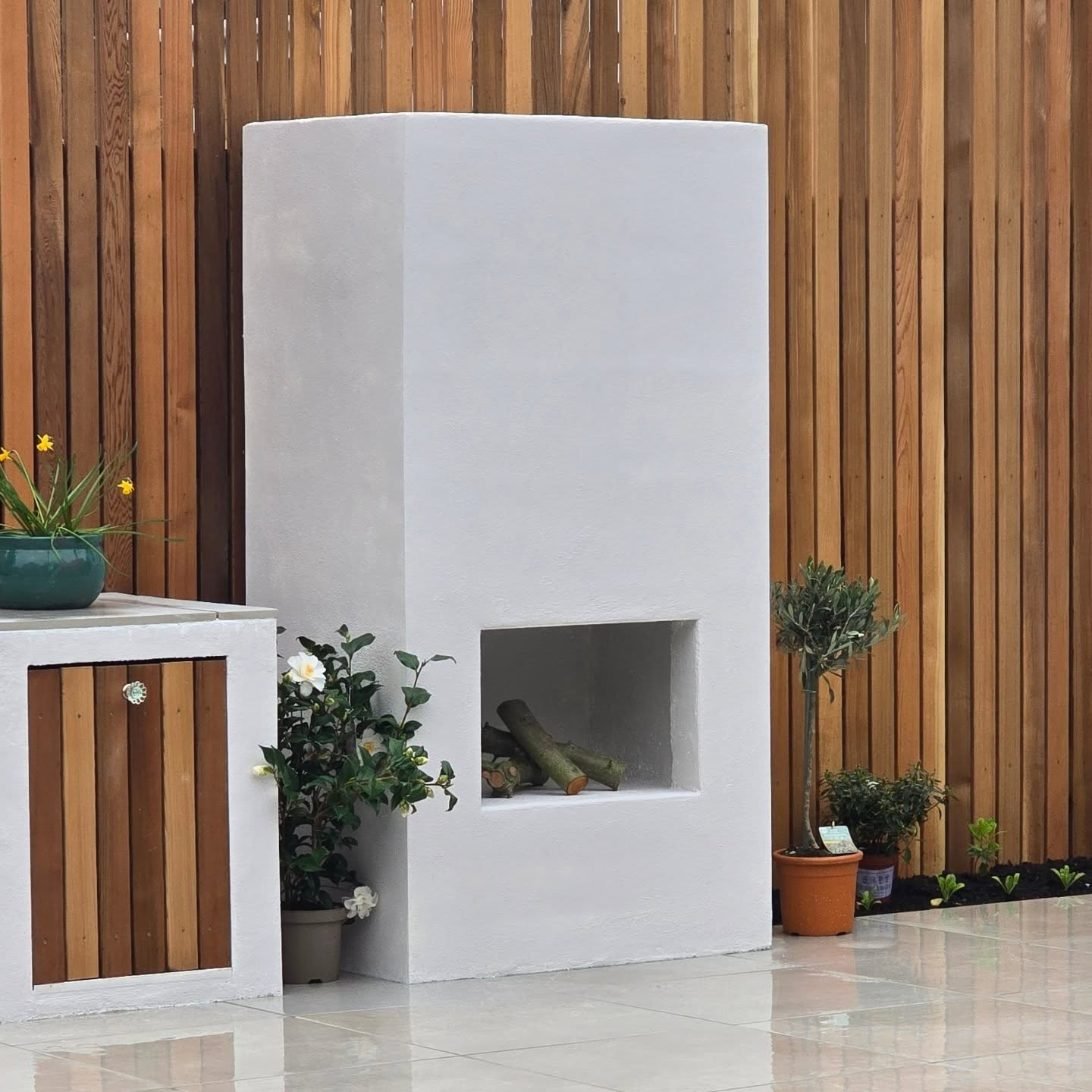 A tall white rendered fireplace with a recessed log store, beside a rendered plinth with a cedar cupboard door, in front of a full-height screen of vertical cedar boards.