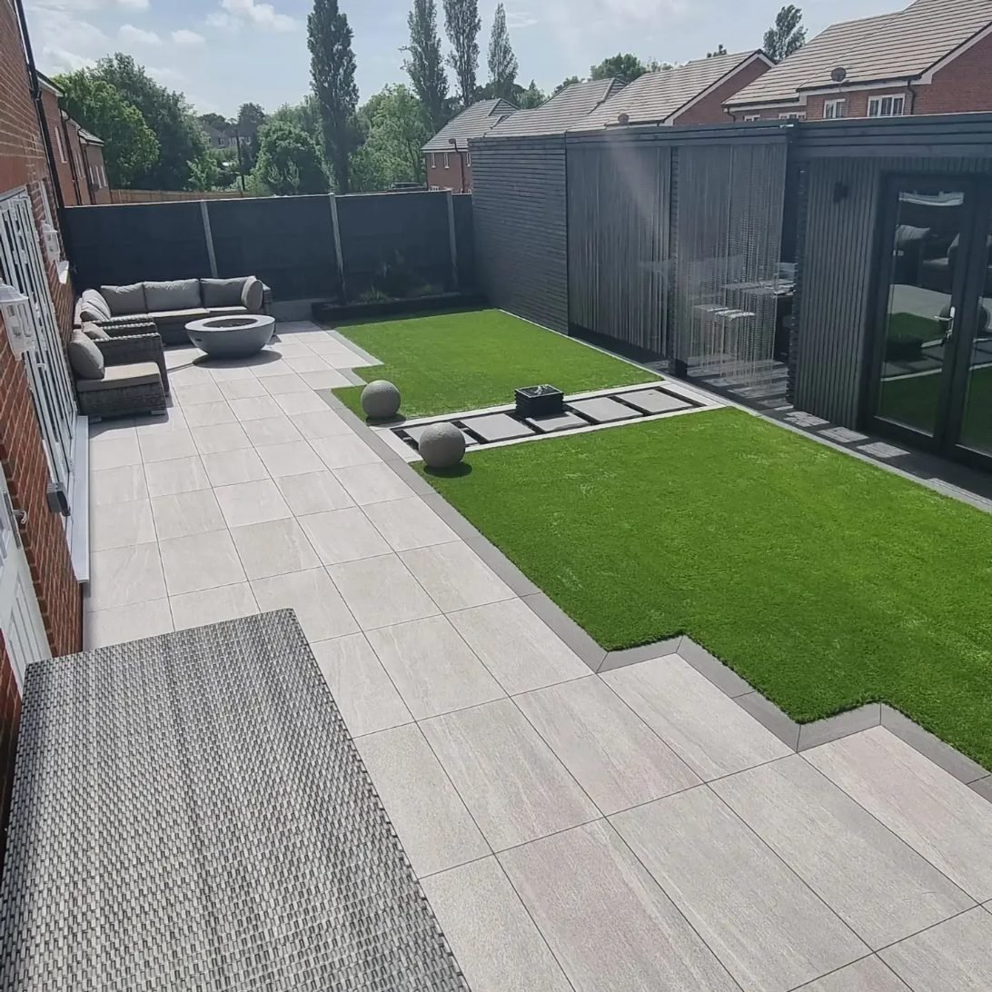 After: a level garden with large pale porcelain paving, two lawns divided by a stepping-stone path, stone spheres, an outdoor sofa and a dark-clad garden room on the right.