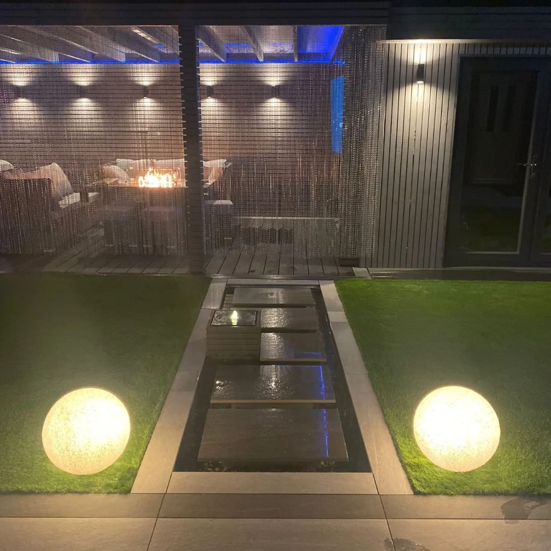 At night, dark porcelain slabs float over a narrow pool between two lawns, flanked by two glowing globe lights, leading to a lit seating area behind a bead curtain.